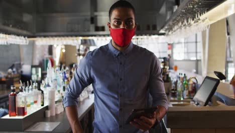 Portrait-of-happy-mixed-race-male-cafe-owner-wearing-face-mask-looking-at-the-camera-and-smiling