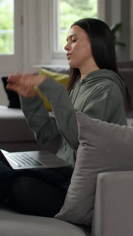 vertical video of woman sitting indoors on sofa working from home using laptop putting hair in ponytail