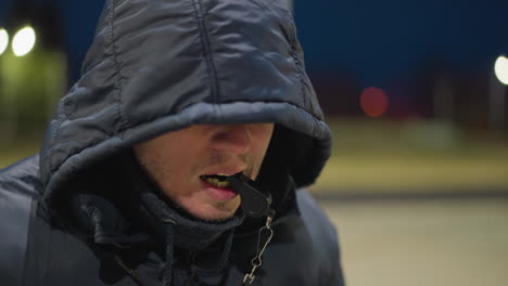 close-up of a man with a hood covering his face, holding a whistle in his mouth, the background is dimly lit, with a blurred view of a building and field