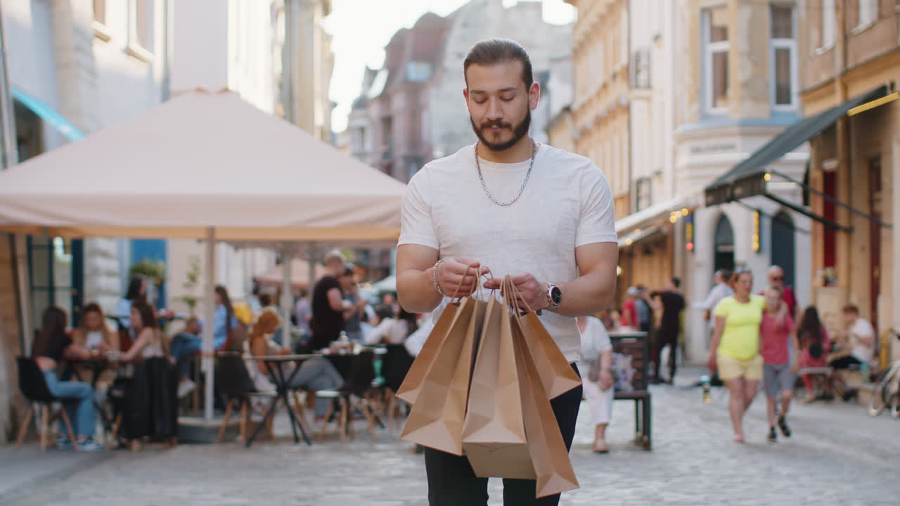 Happy young man guy shopaholic consumer after shopping sale with full bags walking in city street