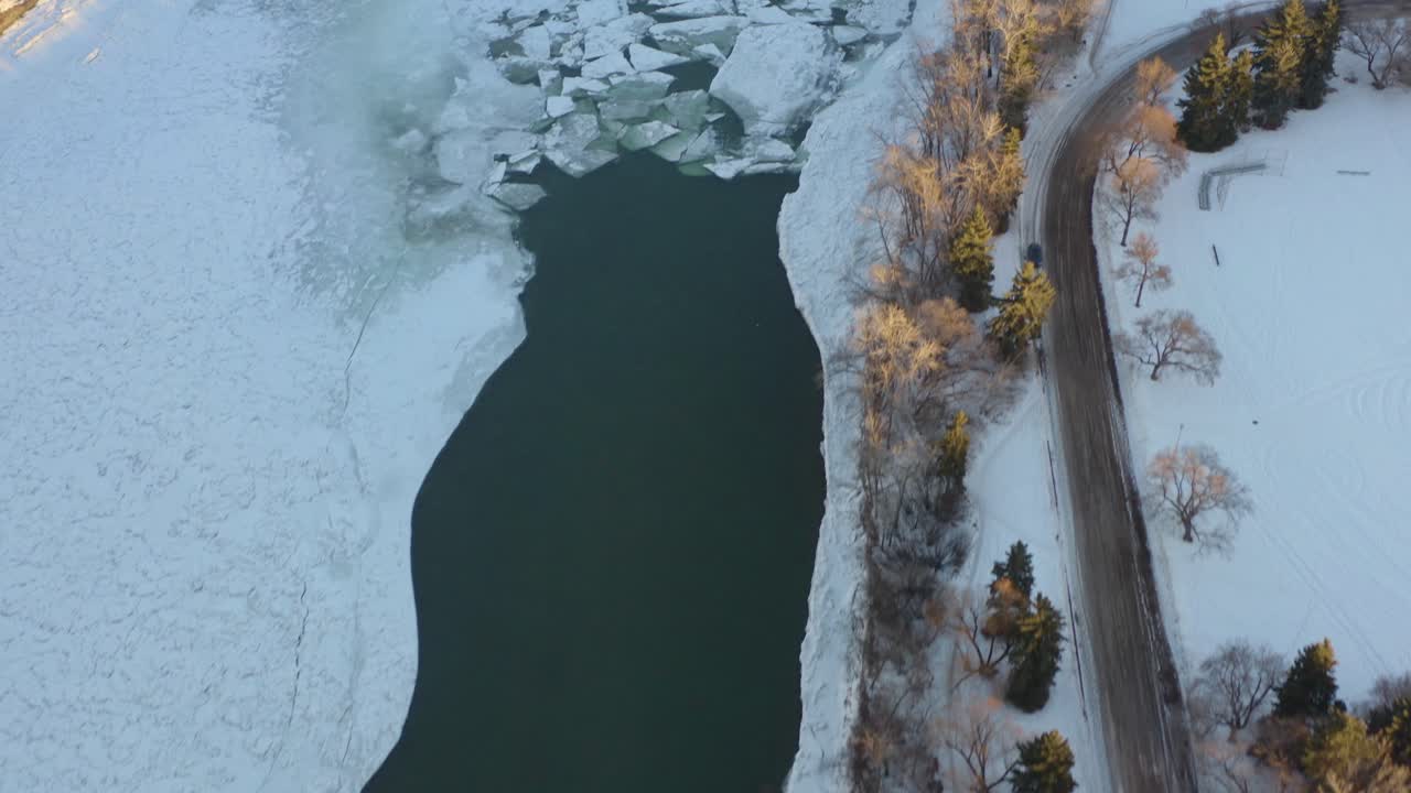 Veduta aerea invernale a volo d'uccello sul fiume North Saskatchewan coperto di neve, ghiaccio e sacche d'acqua fredda, vicino al Kinsmen Park, parte del parco della capitale di Edmonton, in un pomeriggio soleggiato e circondato dalla foresta.