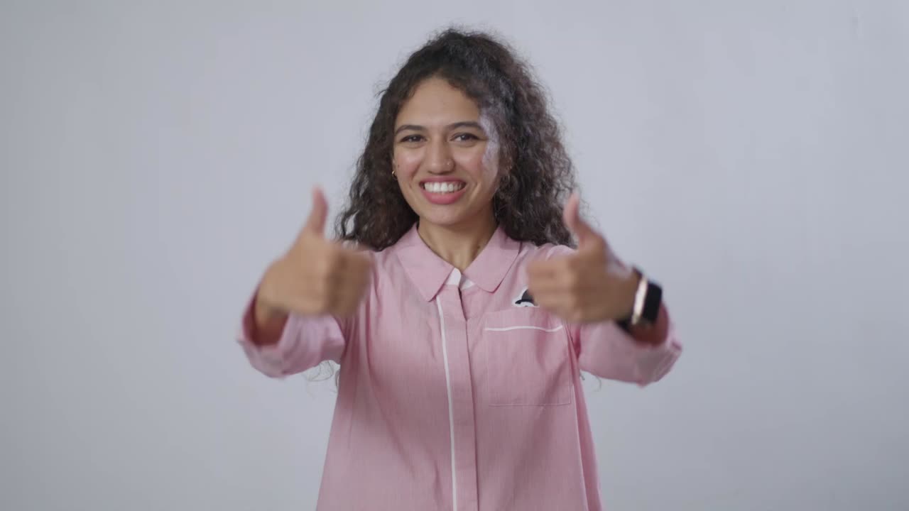 A South Asian woman with a pink shirt reacts with wide-eyed surprise and amazement, expressing astonishment with open hands and touching her face