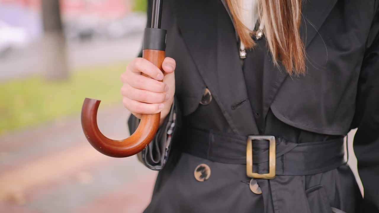 Close-up of woman hand holding wooden handle umbrella while wearing belted black trench coat on rainy autumn day, blurred pavement and greenery in background, stylish seasonal fashion