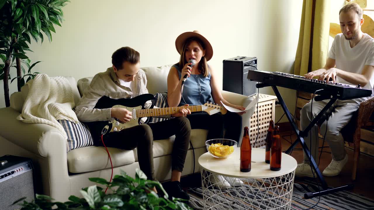 Musical band is rehearsing performance singing and playing musical instruments in home studio looking at music sheet. Bottles and snacks on table are visible.