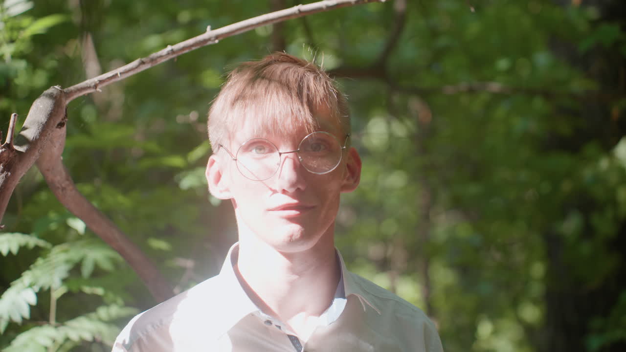 Young man in white shirt and glasses standing in forest observing trees carefully under sunlight, surrounded by dense green foliage, immersed in nature study with quiet thoughtful expression