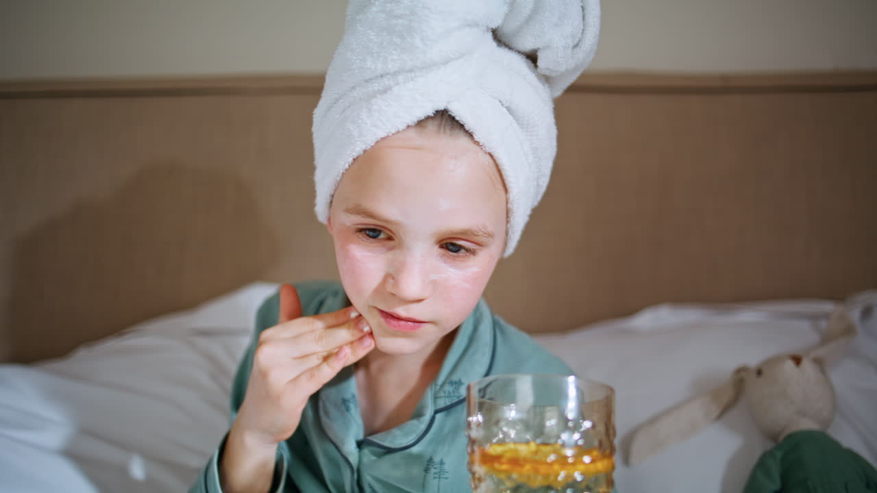 Closeup child moisturizing face with beauty mask at bedroom. Kid holding glass