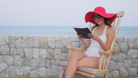 mujer asiática con una hermosa sonrisa está leyendo una revista sentada en una silla de madera cerca de la playa usando monokini blanco y un gran sombrero rojo