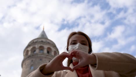 Schönes-Mädchen,-Das-Eine-Medizinische-Schutzmaske-Und-Modische-Kleidung-Trägt,-Formt-Herzform-Mit-Den-Händen-Auf-Dem-Hintergrund-Des-Galata-turms