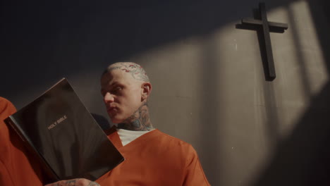 inmate with tattoos and shaved head reading holy bible in prison