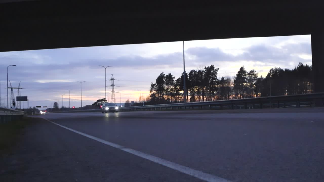 Cars driving to and under a bridge underpass on Pärnu highway during sunset and vehicles drive in slow motion. Trees, lamp posts and electrical lines in the back. White road markings on asphalt