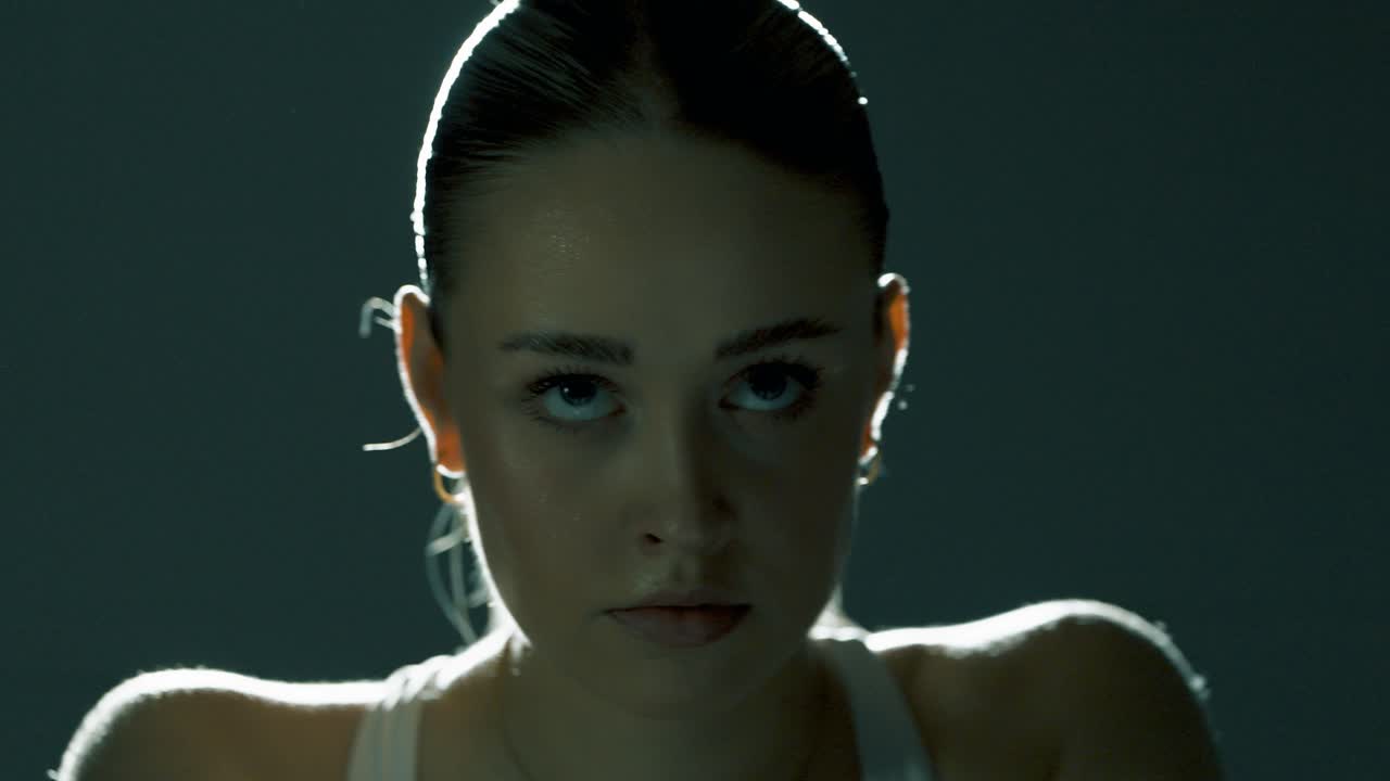 Frontal view of a woman cycling with intense, focused eyes locked forward. The camera glides slightly from left to right as cinematic backlight highlights her sweaty hair and defined shoulders