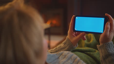 Primer-Plano-De-Una-Mujer-En-Casa-Tumbada-En-El-Salón-Con-Fuego-Transmitiéndose-A-Un-Teléfono-Móvil-Con-Pantalla-Azul