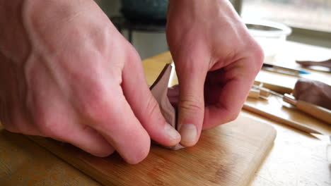 an artist pulling and stretching a piece of soft brown modeling clay and then sculpting with it on a wooden table with tools in an art studio