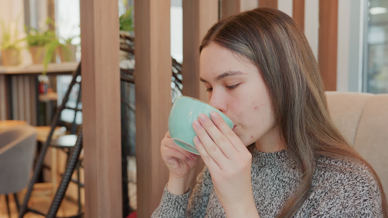 Remote worker drinking cold drink in modern restaurant holds turquoise cup with both hands enjoys refreshing beverage looks around satisfied cozy interior chairs visible with potted plants