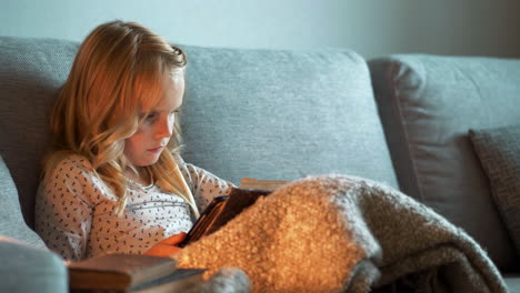 la niña rubia se relaja mientras lee un libro en el sofá por la noche
