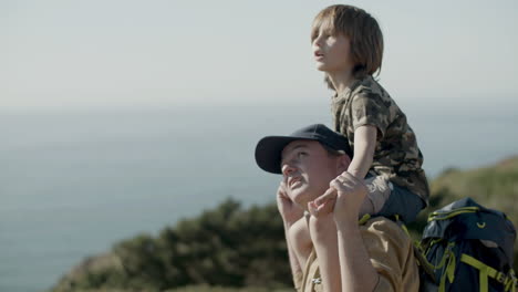 tracking medium shot of father carrying his son on shoulders while hiking in mountains