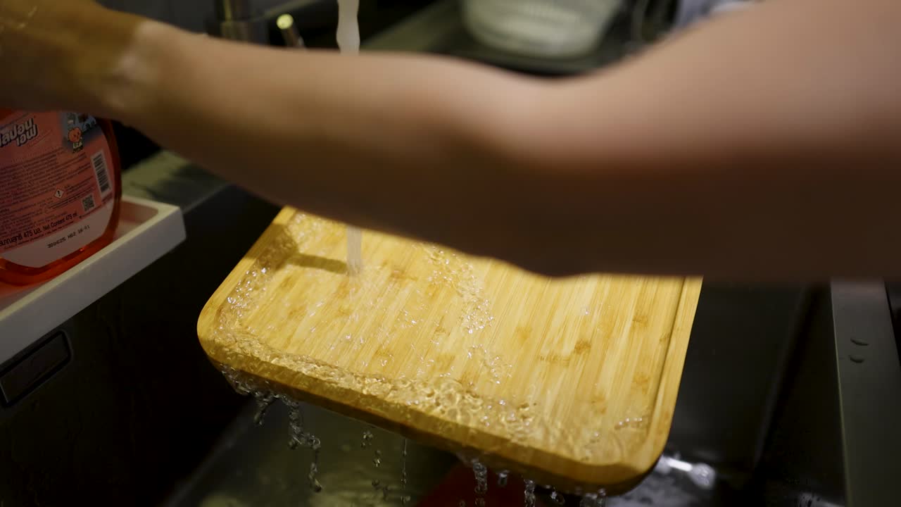 Hands rinse a wooden bamboo chopping board under a faucet in a stainless steel sink