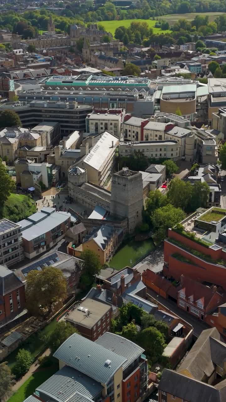 Vertical drone reversing from Oxford Castle and Prison, revealing Oxford’s skyline with historic spires, modern green rooftop architecture, and a blend of ancient and contemporary city landmarks