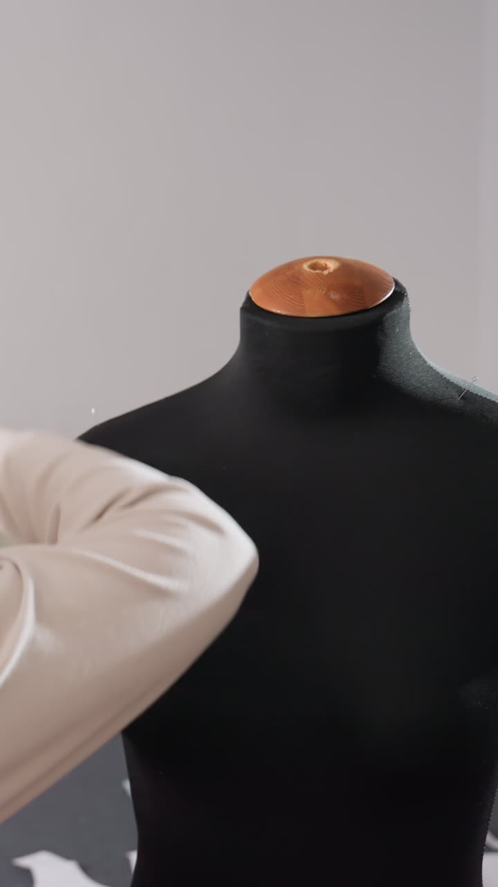 Artesão abotoando colete bege em manequim, Designer inspecionando o alinhamento e a colocação da costura, equipamento de estúdio ao fundo, prova final e avaliação de qualidade, energia criativa focada e refinamento
