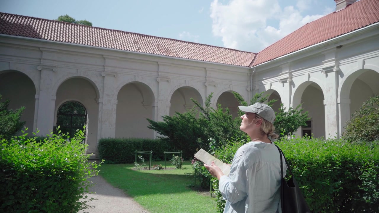 Giovane donna studia una guida turistica cartacea mentre si trova in un soleggiato giardino del chiostro presso il Monastero di Pažaislis.