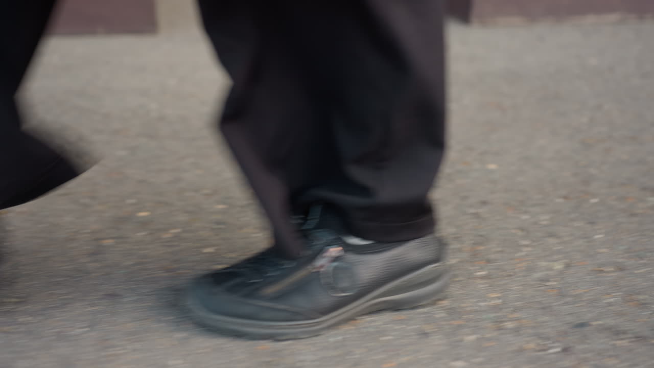 slow motion ground view captures individual in faux leather black shoe stepping along sidewalk as pant hem brushes pavement and steady stride passes camera highlighting texture and movement