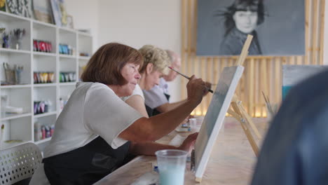 Vista-Lateral-De-Una-Gente-Mayor-Feliz-Sonriendo-Mientras-Dibuja-Como-Actividad-Recreativa-O-Terapia-En-Clase-De-Pintura-Junto-Con-El-Grupo-De-Mujeres-Y-Hombres-Jubilados