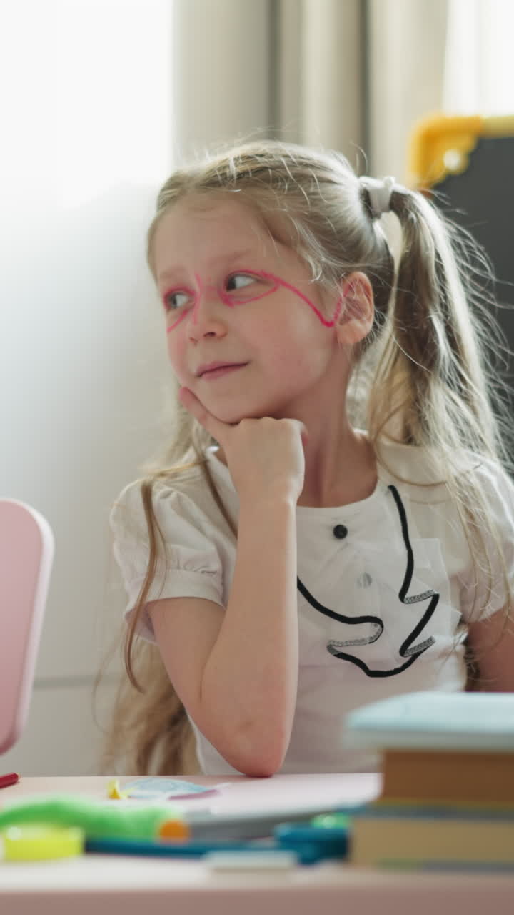 Little boy and girl look at each other and nod puffing out cheeks. Couple of children with painted glasses laughs carelessly sitting at desk in light classroom