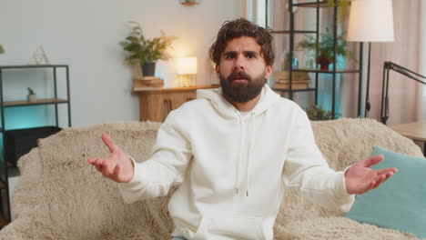 young indian man raising hands in indignant expression asking reason of failure at home apartment