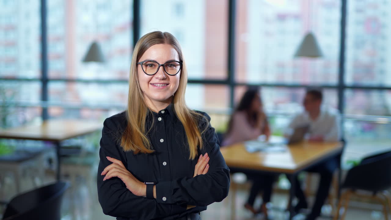 Smiling lady in braces puts on glasses. Confident young woman at workplace. Suddenly she feels headache and touches her temples. People in blur at backdrop.