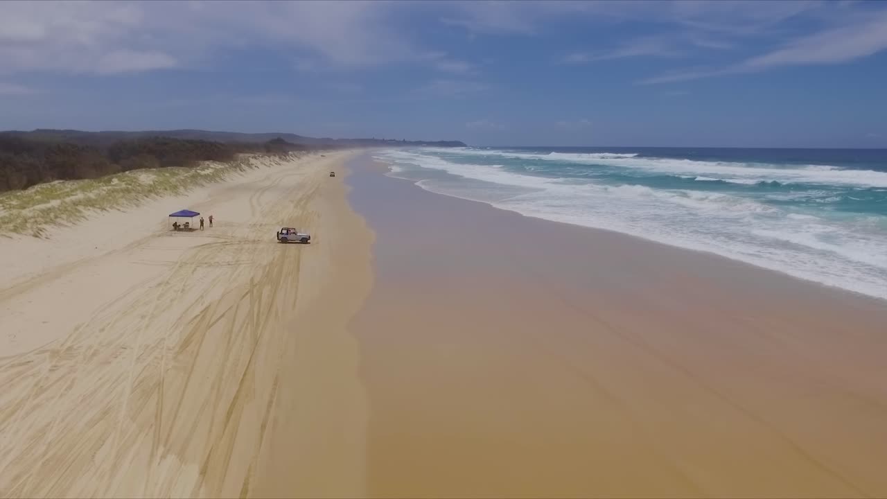 Low aerial drone shot travelling North up Main Beach on Queensland's North Stradbroke Island towards a camp site, with 4x4 four wheel drive vehicles driving on the beach