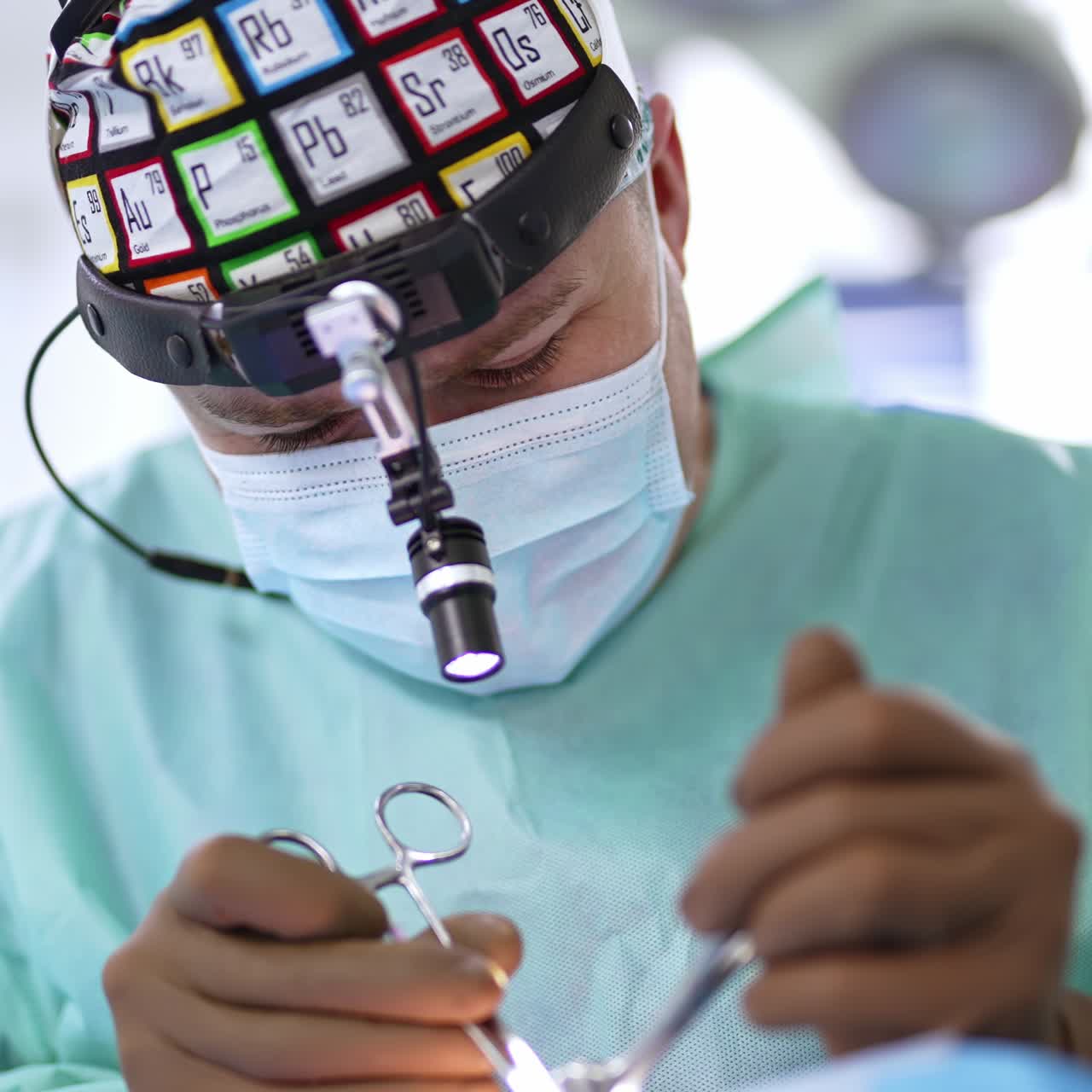Un chirurgo sorridente con mascherina e dispositivo sulla testa prende degli strumenti di metallo. Le mani inguantate dell'assistente passano gli strumenti al dottore. Primo piano