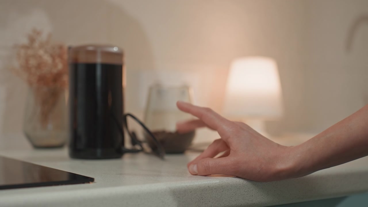 Main sur le comptoir de la cuisine près d'une enceinte, lueur chaude d'une lampe, appareil filaire et verre en arrière-plan, les doigts planent puis pressent le bord du comptoir, amateur de musique nocturne tendant la main vers un appareil audio, ambiance intime et douillette