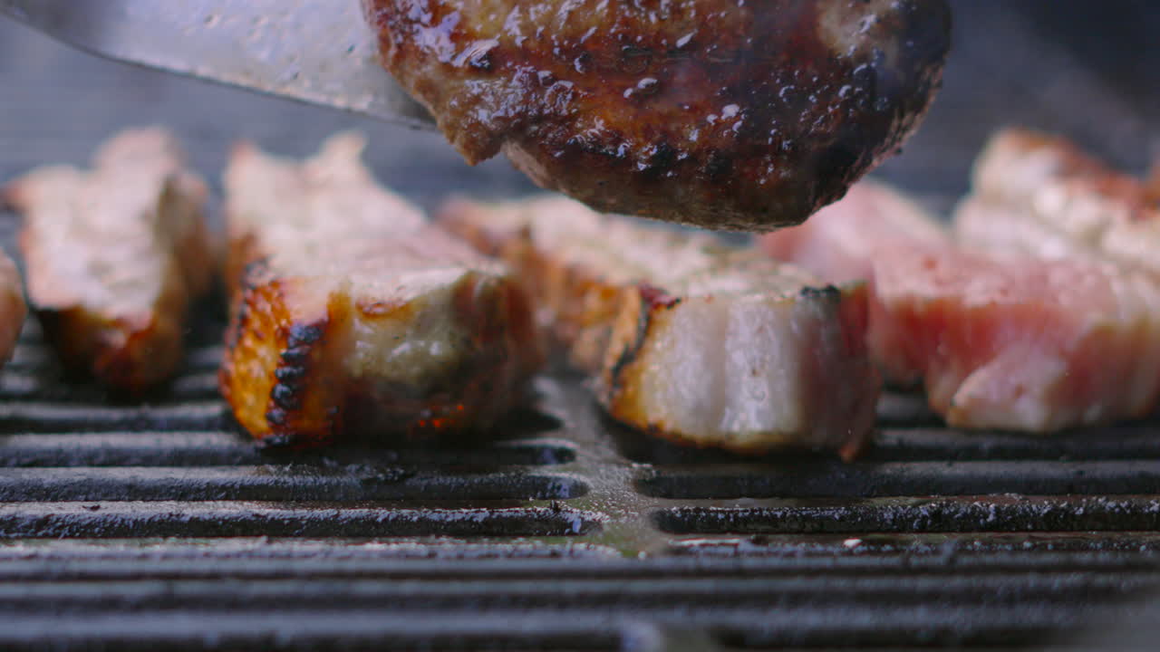 Beef Burger Being Flipped on Grill with Fire Flames Engulfing Hamburger and Flaring Towards Camera in Slow Motion. High Quality Close Up Barbecue Summer Cooking Food. Pork Belly Slices in Background.