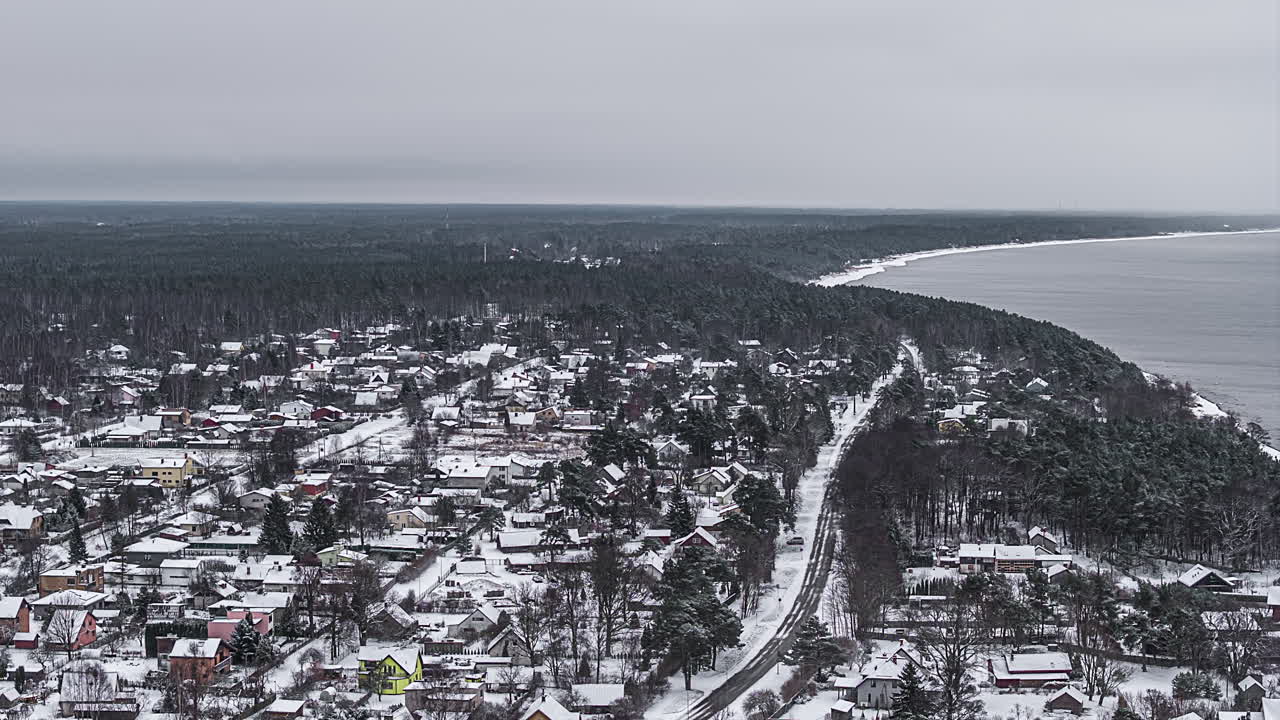 Subtil drone-timelapse over en snødekt kystby og furuskog, som følger en svingete vei mot sjøkysten under myke grå vinter skyer