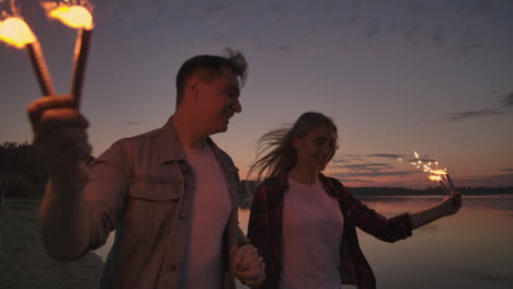 Young-lovers-man-and-woman-cheerfully-run-along-the-beach-with-hot-sparkling-rot-in-their-hands-in-slow-motion.-Romantic-relationship.-Sparklers-burn-in-the-hands-of-lovers