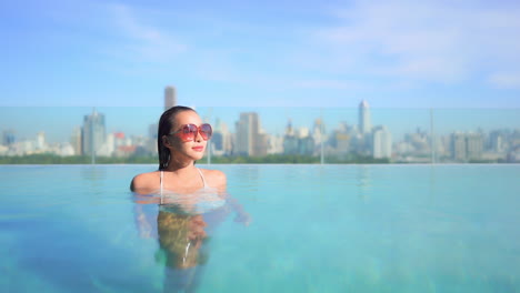 Mujer-Asiática-Sexy-En-La-Piscina-Infinita-En-La-Azotea-Con-El-Paisaje-Urbano-De-Fondo