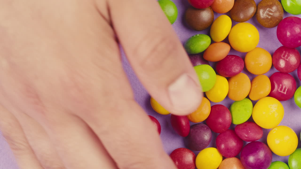 Colorful dragee candies on purple background. Woman hands move the bonbons. Summer bright studio shot for store, supermarket, close up top view. Children sweets made of sugar, mix of lollipops.