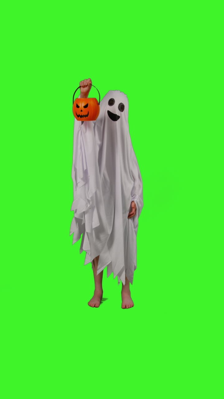 vertikales video in voller länge von einem kind, das sich in ein geisterkostüm verkleidet hat, das an halloween trick oder treat macht und einen kürbischförmigen jack-o-lantern-eimer gegen den grünen bildschirm des studios hält