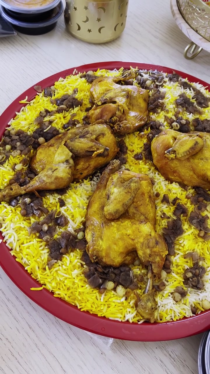 Close-up sudut tinggi hidangan machboos ayam tradisional Kuwait di atas meja makan kayu. Nampan merah besar itu menyajikan nasi kuning, ayam panggang, dan bawang hashwu dalam suasana dalam ruangan yang cerah