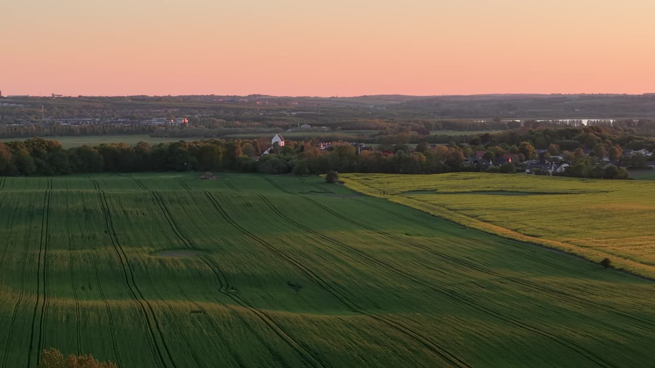 Aerial drone view of green farmland fields at sunset with a rural town and distant horizon
