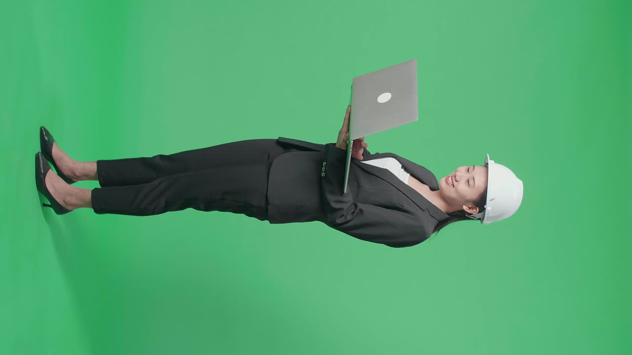 Full Body Side View Of Smiling Asian Female Engineer With Safety Helmet Typing On A Laptop In The Green Screen Studio