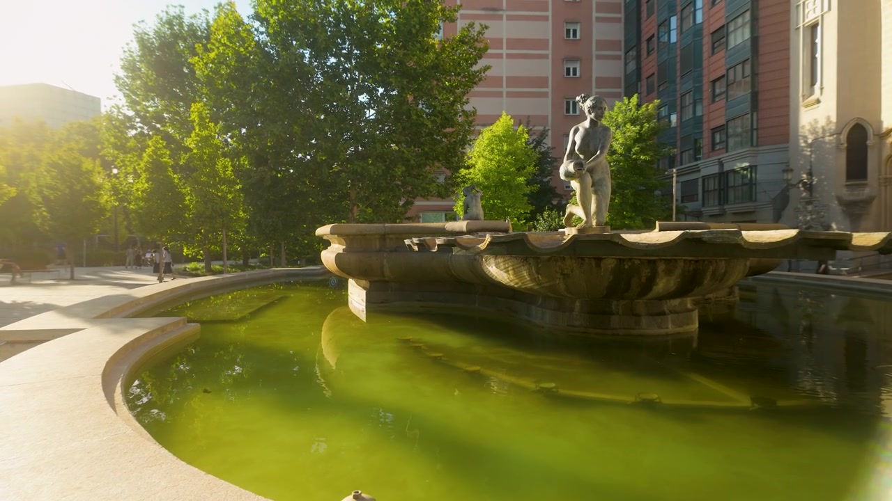Una serena fuente de piedra con una estatua femenina en una plaza de Madrid, rodeada de exuberantes árboles verdes y arquitectura urbana durante un cálido atardecer.