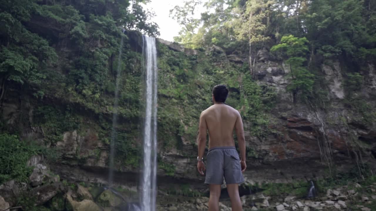 A man, viewed from behind, stands on a rock platform at the base of the Misol-Há waterfall in Chiapas, México