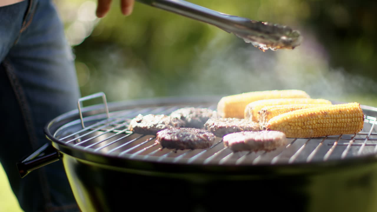 Grilling burgers and corn on barbecue grill, african american man using tongs for cooking, outdoors
