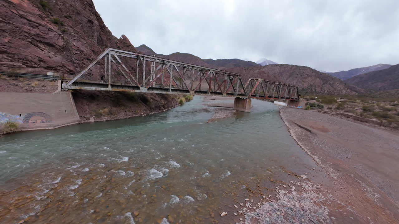 Dynamic FPV drone performs sharp left turn, flying directly under historic railway bridge on a cloudy day, river underneath