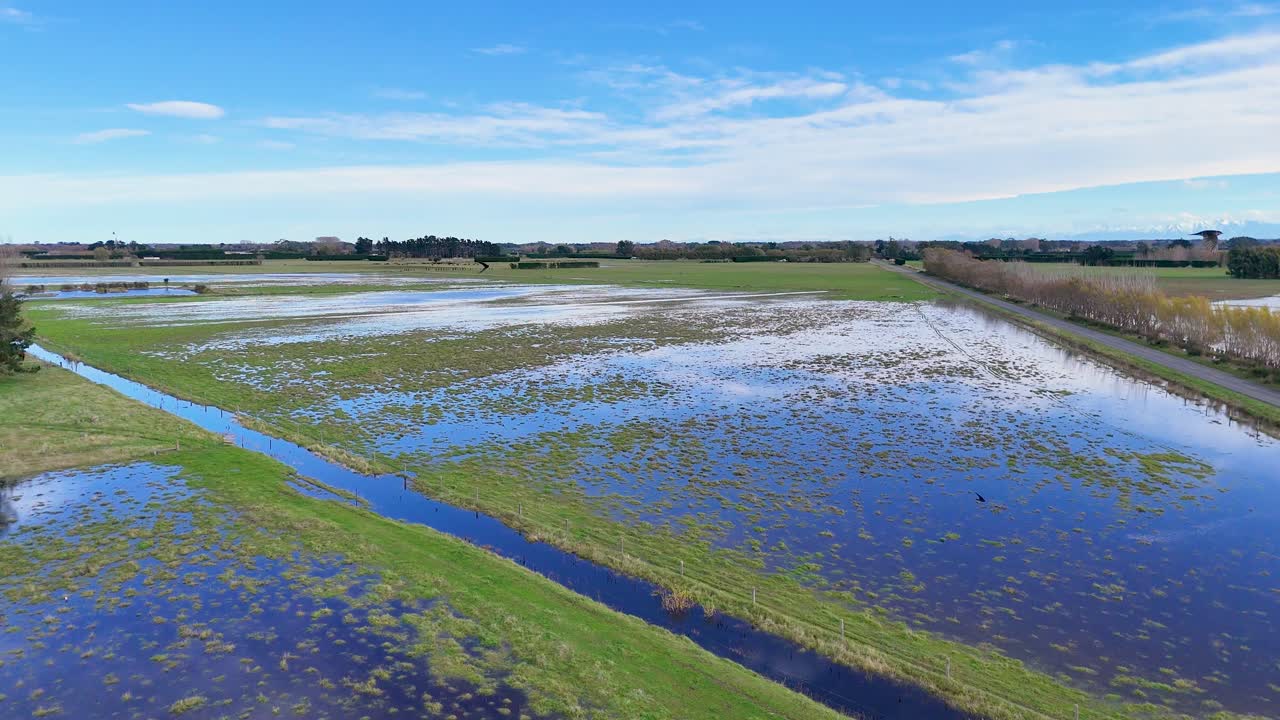 Aerial footage of flooded fields in Christchurch, New Zealand. Bright daylight illuminates the expansive waterlogged landscape with clear blue skies