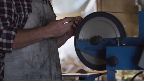 midsection of caucasian male knife maker in workshop wearing glasses and using sander