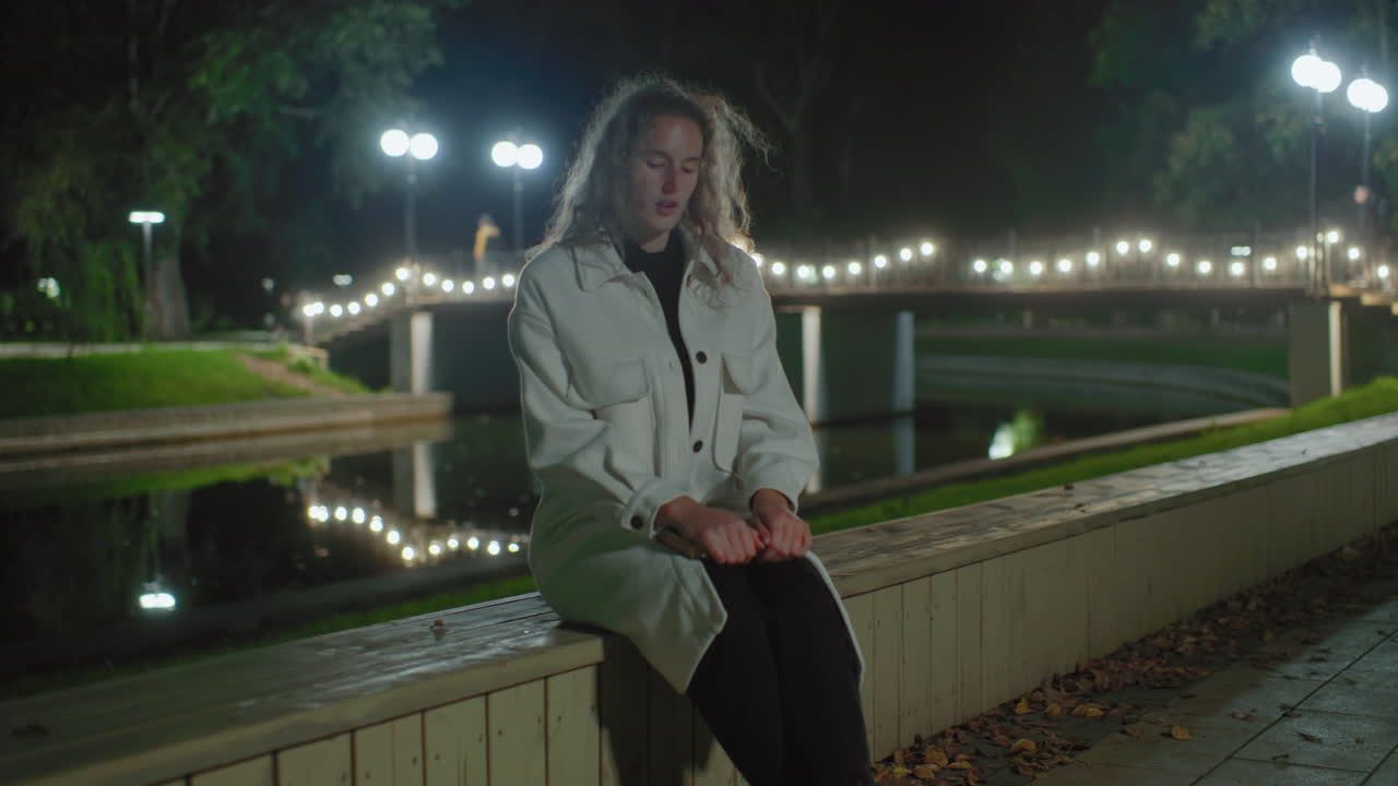 Girl walks up to wooden fence beside calm water at night, sits down, places book on leg, casually reading while people stroll over lit bridge in background, gentle reflections and fallen leaves