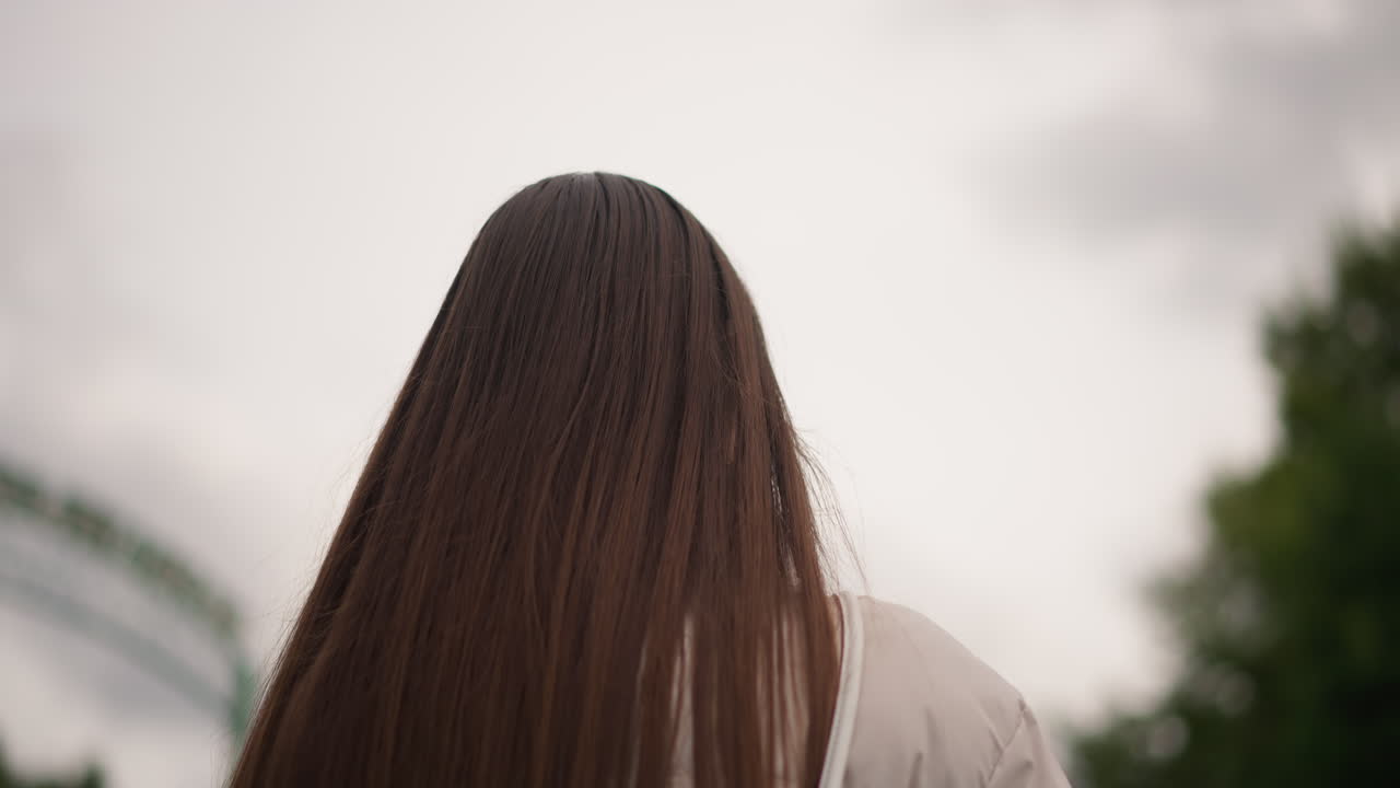 Giovane donna con trecce in piedi all'aperto vicino all'entrata di un parco sotto un cielo nuvoloso, movimento lento della testa, umore contemplativo, una leggera brezza solleva le trecce, toni cinematografici tenui e pacata solitudine