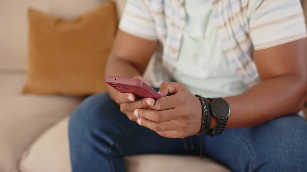 Afroameričan v pruhovaném tričku ťuká a posouvá červený telefon na gauči po zprávě, usmívá se.